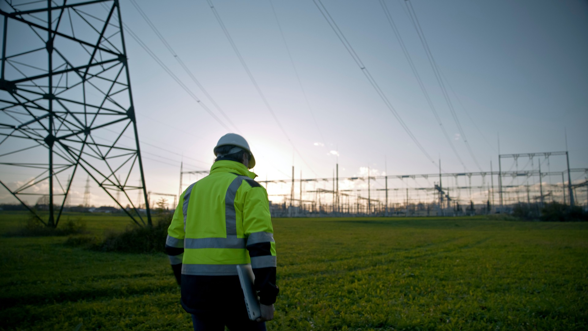 Rear View Of Male Electrical Engineer In Reflective Clothing While Walking At Power Station During Sunset